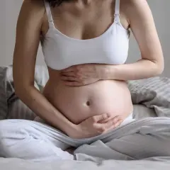 Gravid kvinna sitter på sängen med händerna på magen. Hon känner sig lite dålig i magen, en vanlig känsla under graviditeten.