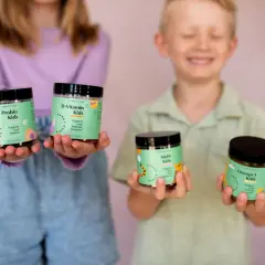 Två barn håller upp burkar med vitaminer. En flicka håller Probio Kids och D-Vitamin Kids, medan en pojke håller Multi Kids och Omega 3 Kids. Perfekt för vitamin barn!