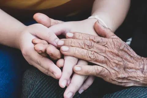 Två händer, en ung och en gammal, flätas samman. Den äldre handen visar tydliga tecken på biologisk ålder, medan den yngre är slät och mjuk. En symbol för livets gång och generationer.