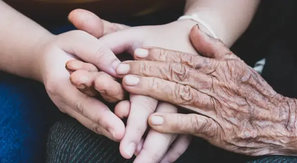 Två händer, en ung och en gammal, flätas samman. Den äldre handen visar tydliga tecken på biologisk ålder, medan den yngre är slät och mjuk. En symbol för livets gång och generationer.