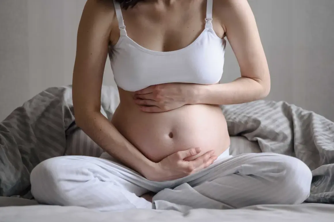 Gravid kvinna sitter på sängen med händerna på magen. Hon känner sig lite dålig i magen, en vanlig känsla under graviditeten.