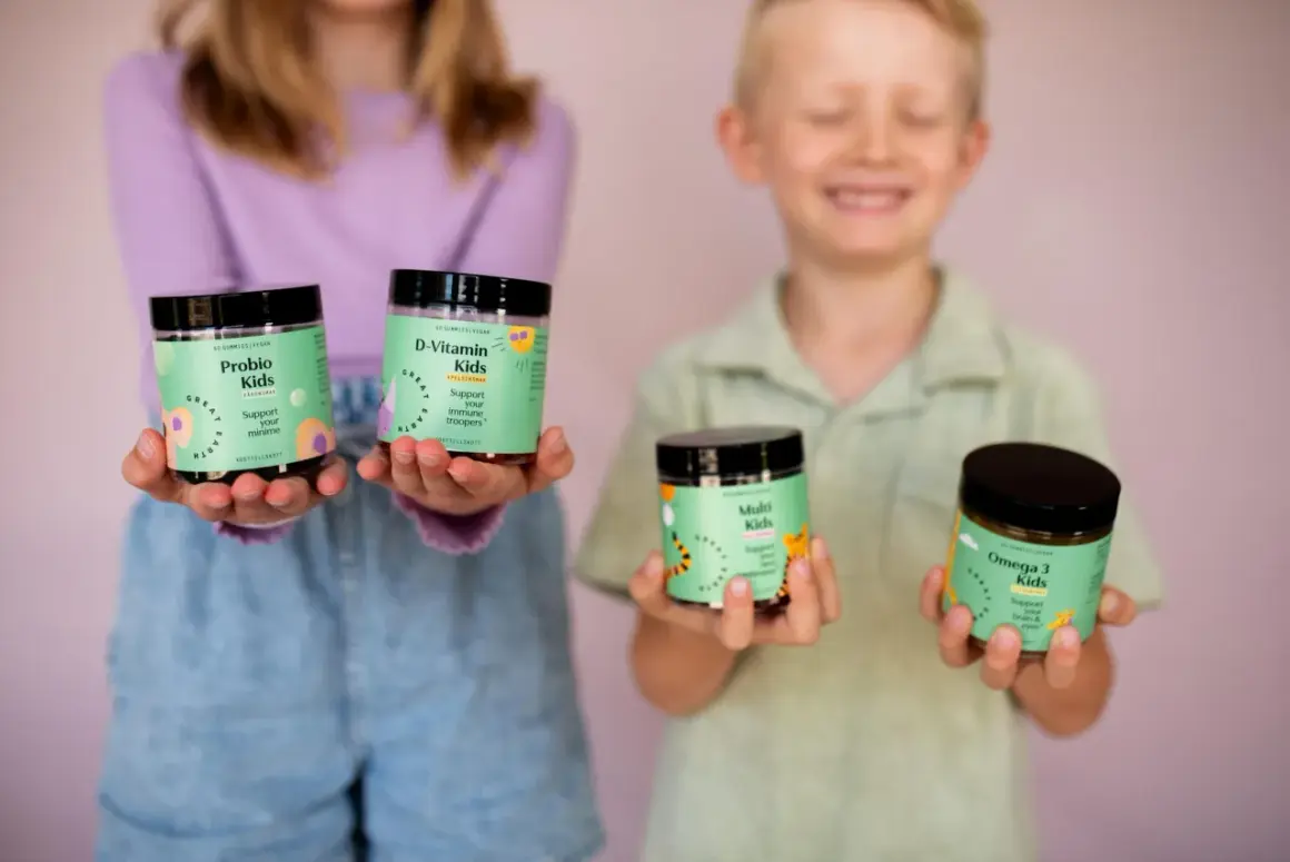 Två barn håller upp burkar med vitaminer. En flicka håller Probio Kids och D-Vitamin Kids, medan en pojke håller Multi Kids och Omega 3 Kids. Perfekt för vitamin barn!