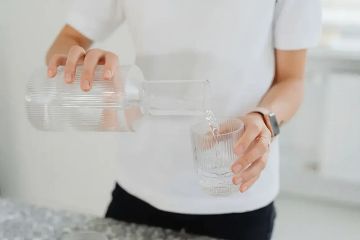 En person häller upp vatten från en glasflaska till ett glas. En hälsosam dryck som mot rethosta.