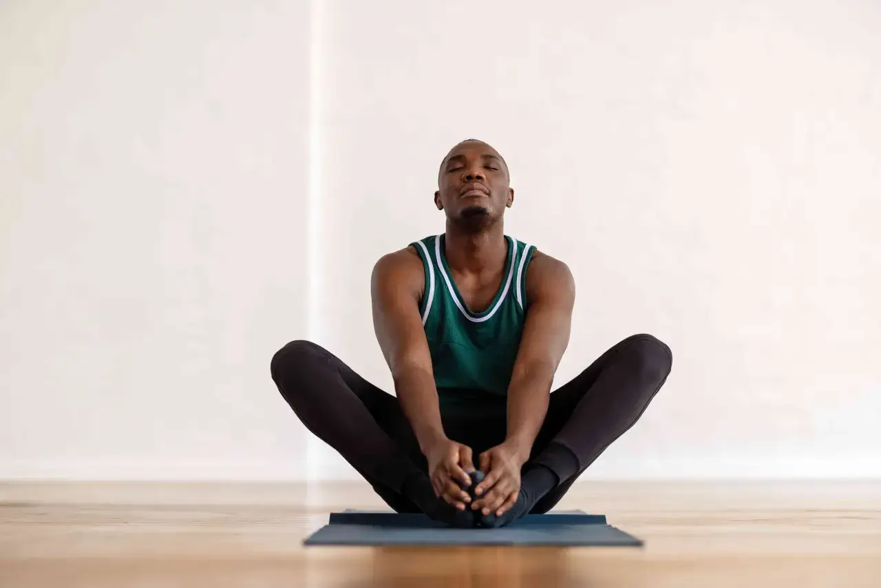 Man i gr&ouml;n linne och svarta tr&auml;ningskl&auml;der sitter p&aring; en yogamatta i fj&auml;rilsposition, med slutna &ouml;gon. En avslappnande st&auml;llning som kan hj&auml;lpa till att f&aring; ut gaser ur magen snabbt.