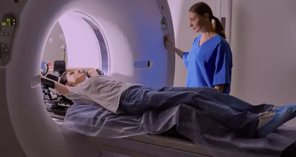 En patient ligger på en brits i en MR-scanner, medan en sjuksköterska justerar inställningarna. Mr Ryggs undersökning pågår.