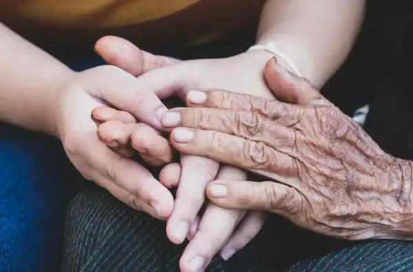 Två händer, en ung och en gammal, flätas samman. Den äldre handen visar tydliga tecken på biologisk ålder, medan den yngre är slät och mjuk. En symbol för livets gång och generationer.