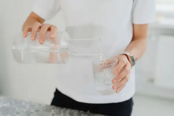 En person häller upp vatten från en glasflaska till ett glas. En hälsosam dryck som mot rethosta.